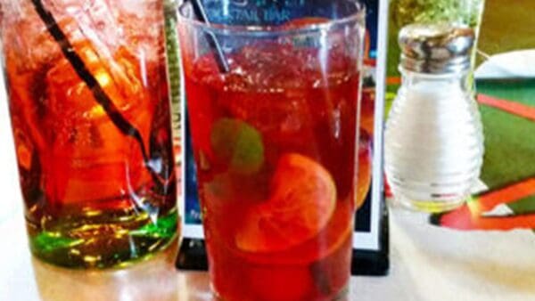 Two glasses with ice and red beverages on a table. One drink has fruit slices inside. There is also a salt shaker and a menu in the background.