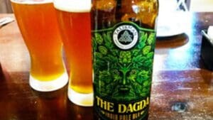 A bottle of "The Dagda Warrior Pale Ale" is on a wooden table, with two filled beer glasses in the background. The label features a green design with an intricate figure.