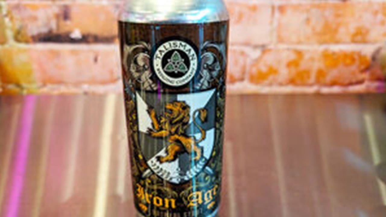 A can of AleSmith Brewing Company's Iron Age beer with a lion crest design stands on a metallic surface against a brick wall background.