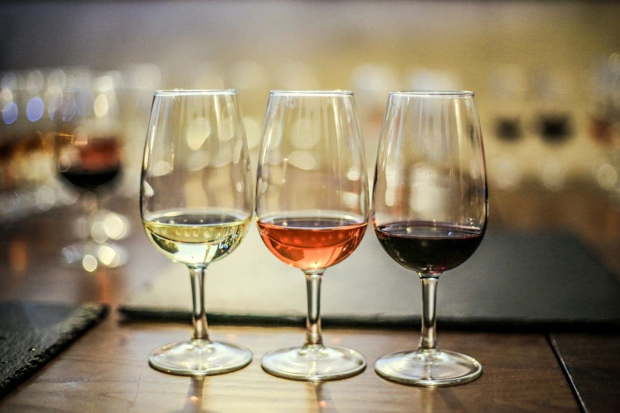 Three wine glasses with different types of wine (white, rosé, and red) placed in a row on a wooden table. Background shows more out-of-focus glasses and a soft lighting.